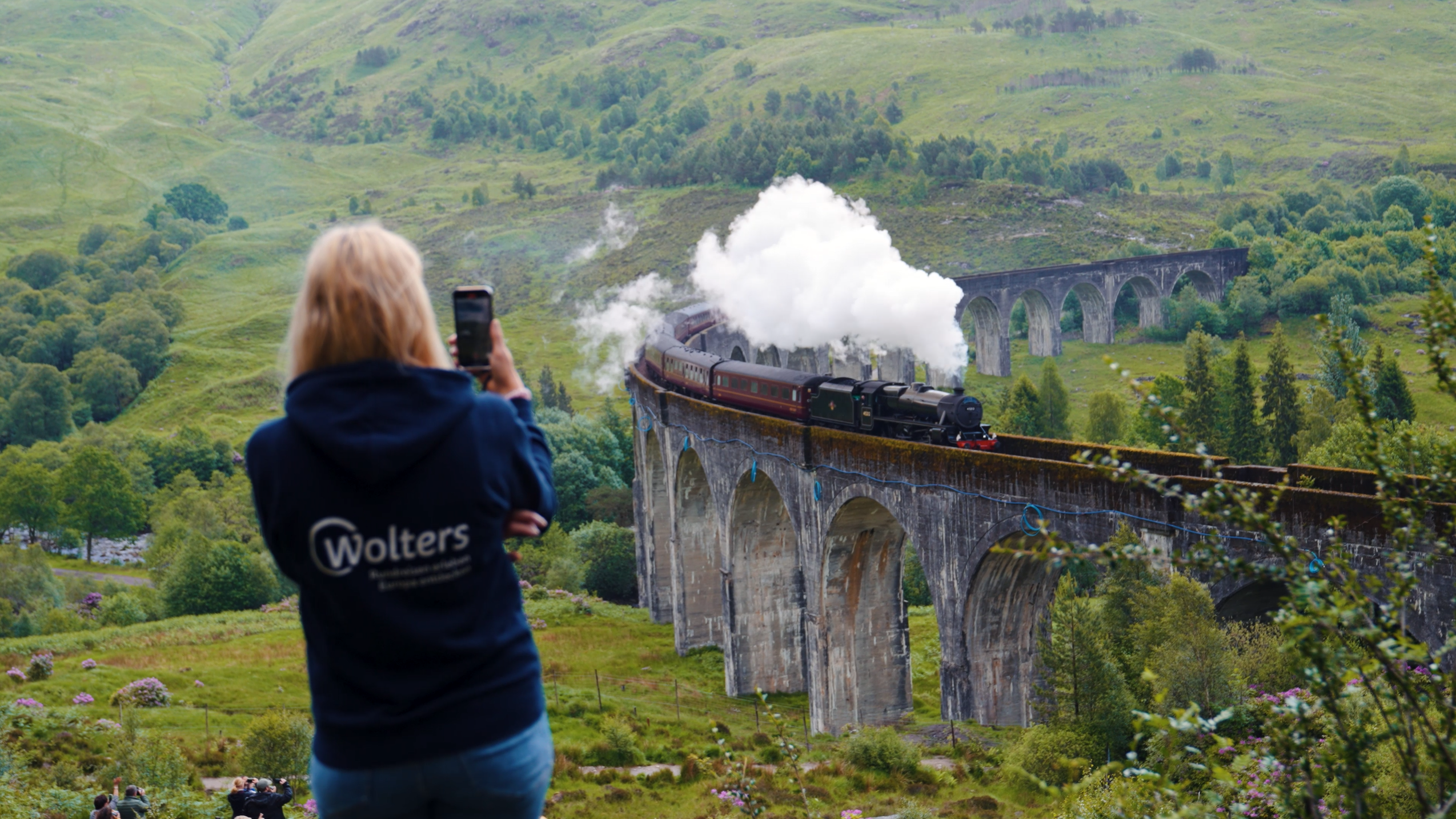 Blonde Wolters Mitarbeiterin vor fotografiert den Jacobite Steam Train auf dem Glenfinnan Viadukt Eine Person fotografiert den berühmten Dampfzug, der über das Glenfinnan Viadukt in den schottischen Highlands fährt. Umgeben von grünen Hügeln und dichter Natur vermittelt die Szene den Zauber klassischer Zugreisen und unvergesslicher Schottland-Erlebnisse.