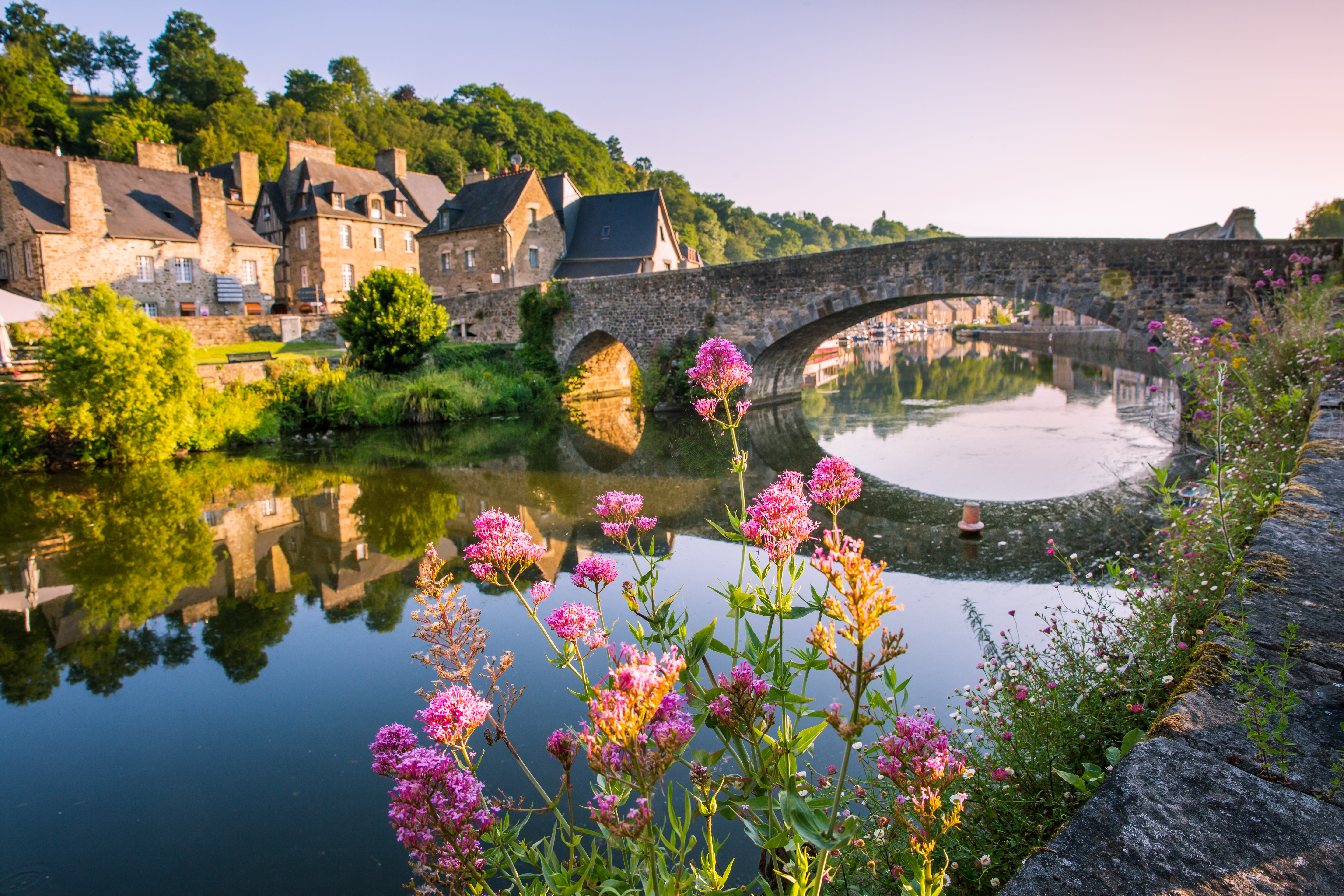 Malerische Steinbrücke in Dinan in der Bretagne, gesäumt von historischen Fachwerkhäusern am Ufer des Flusses Rance mit bunten Blumen im Vordergrund.