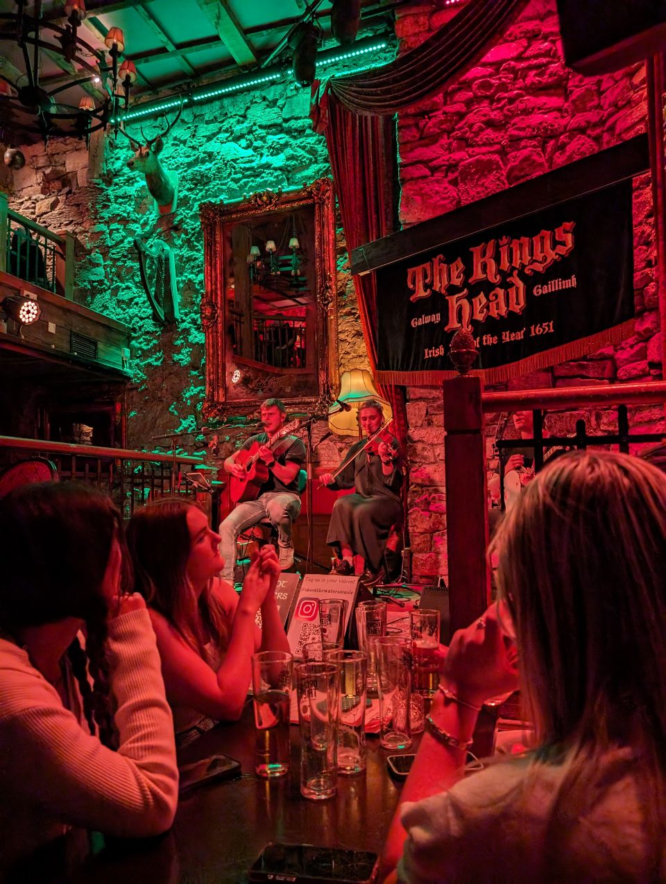 Auf dem Bild ist eine Live-Musik-Session im berühmten Pub „The King's Head“ in Galway, Irland, zu sehen. Zwei Musiker spielen für die Gäste, während die stimmungsvolle rote und grüne Beleuchtung die alten Steinmauern des Pubs hervorhebt und eine authentische Atmosphäre schafft.