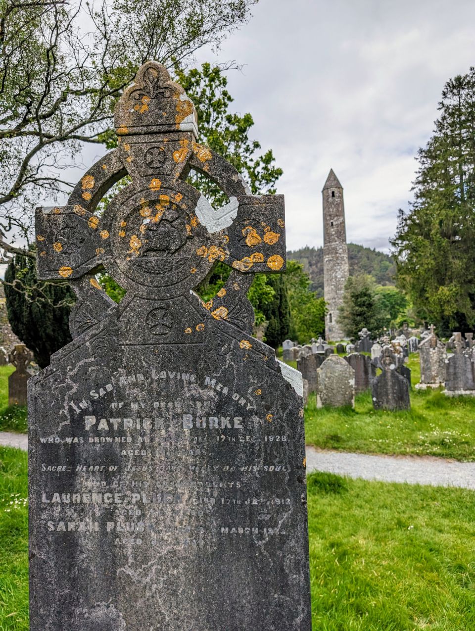 Ein altes keltisches Kreuz dominiert den Vordergrund dieses Bildes, aufgenommen auf dem historischen Friedhof von Glendalough in Irland. Im Hintergrund ragt der berühmte Rundturm der Klostersiedlung in den Himmel und vervollständigt diese geschichtsträchtige und atmosphärische Szene.