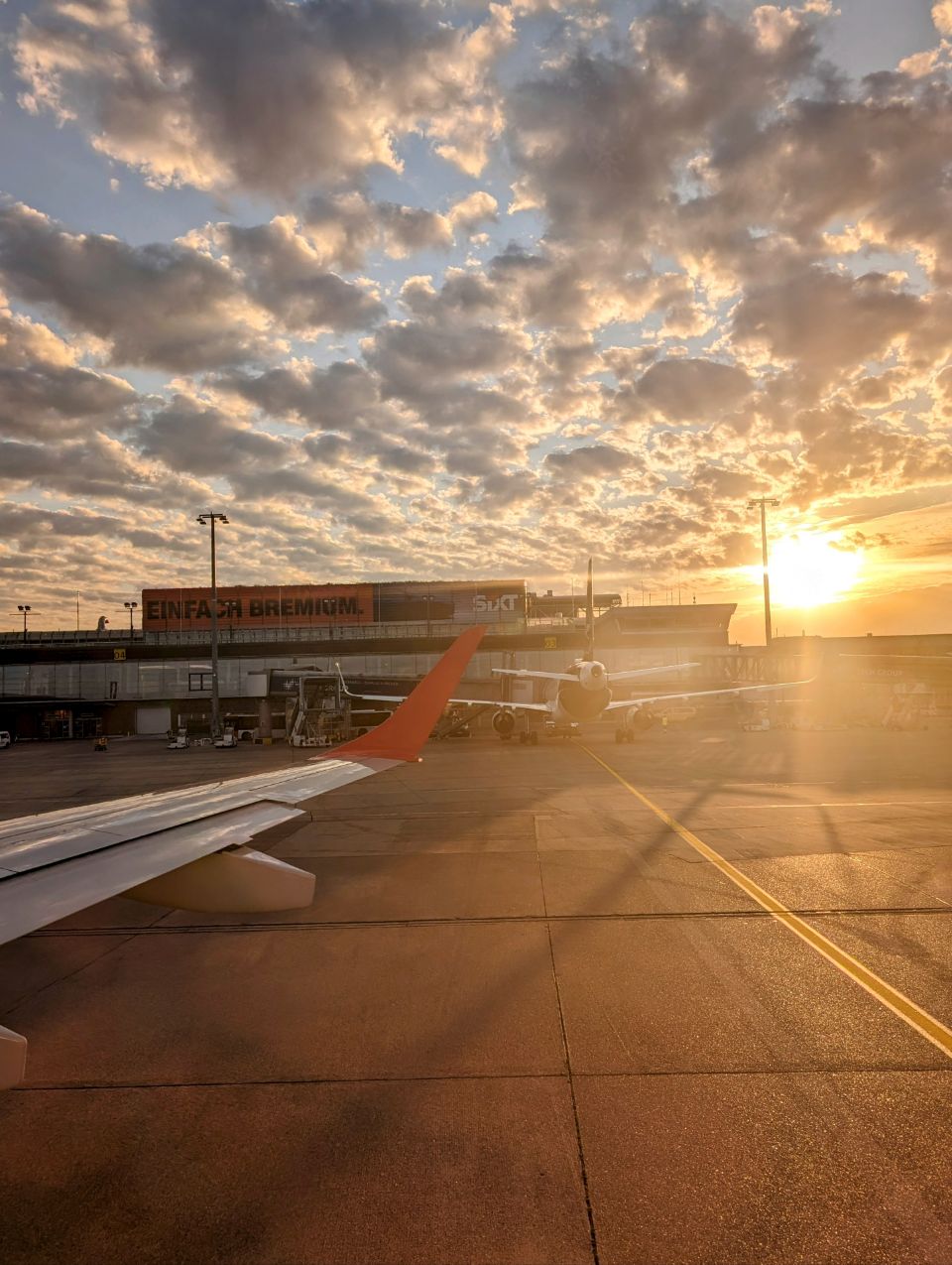 Der Blick aus dem Flugzeugfenster zeigt den Flughafen Bremen in einer stimmungsvollen Sonnenuntergangs-Szene. Man erkennt die in warmes Licht getauchte Flugzeugtragfläche und das Terminalgebäude, was die besondere Atmosphäre kurz vor einem Start in den Abendhimmel einfängt.