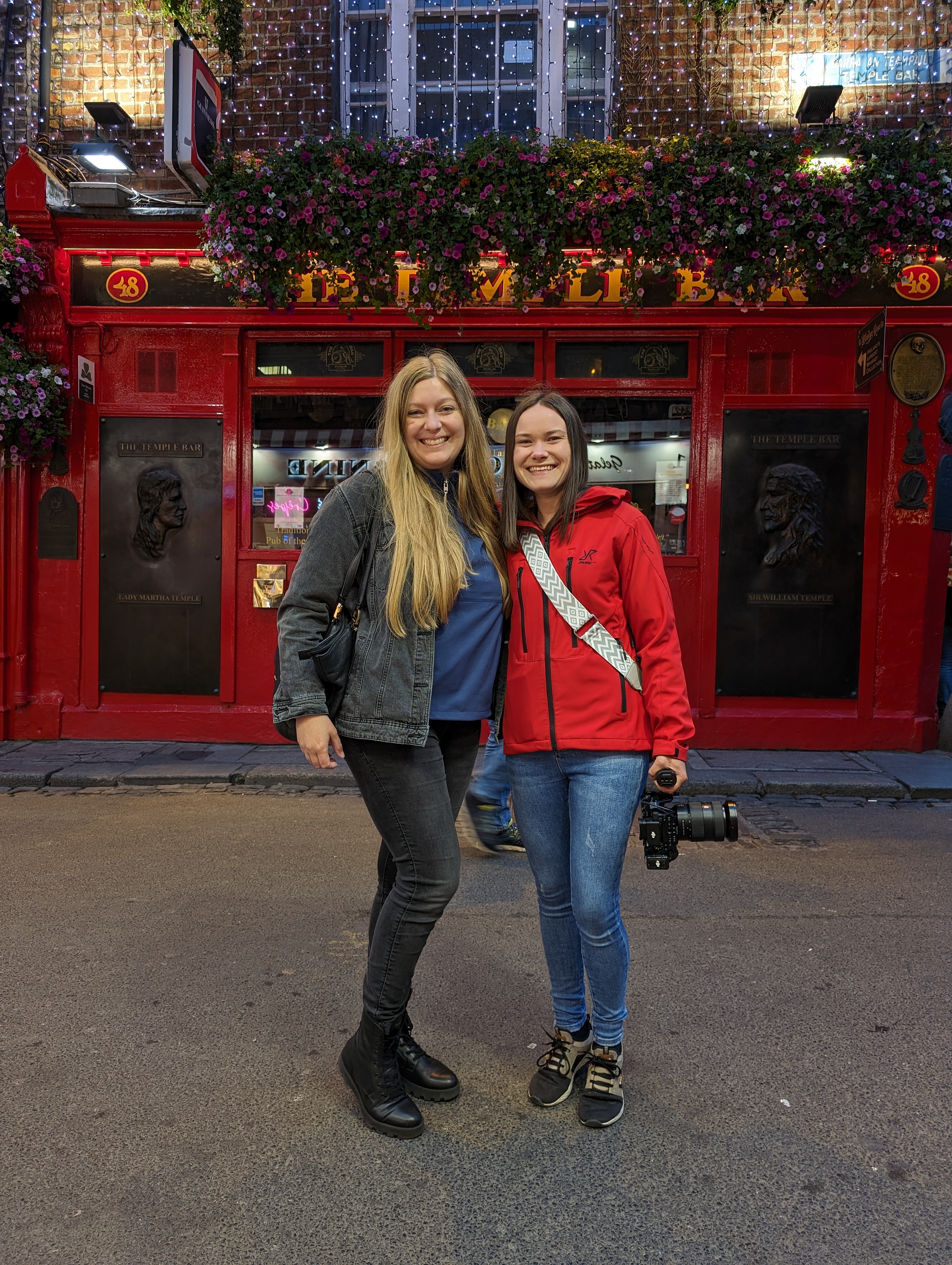 Vor der ikonischen roten Fassade des weltberühmten „The Temple Bar“ Pubs in Dublin posieren hier zwei Frauen für ein Erinnerungsfoto. Die nächtliche Beleuchtung und die fröhliche Stimmung der beiden fangen die lebendige Atmosphäre dieses beliebten touristischen Hotspots perfekt ein.