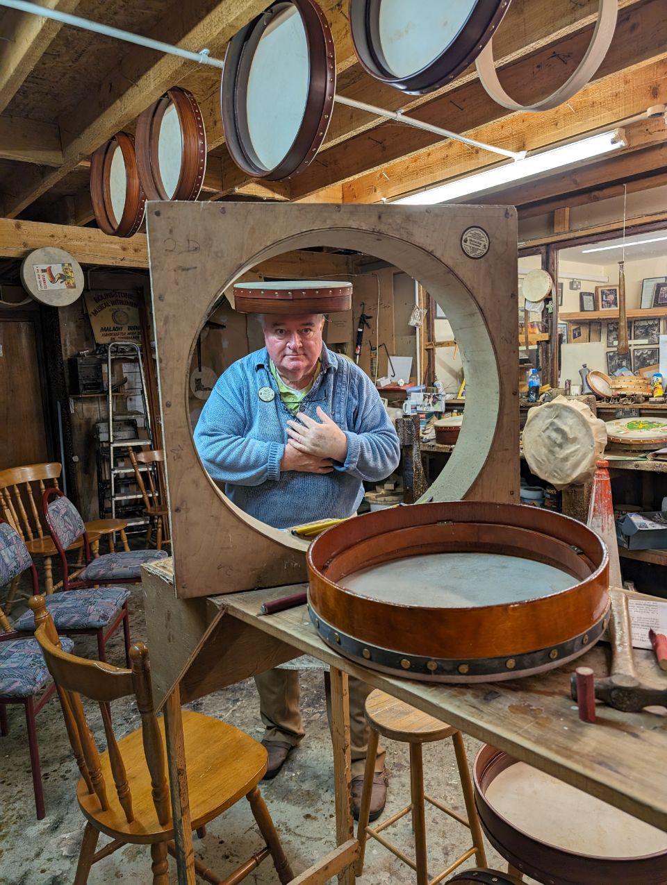 Auf dem Bild ist der berühmte Bodhrán-Macher Malachy Kearns in seiner Werkstatt in Roundstone, Irland, zu sehen. Umgeben von seinen handgefertigten irischen Rahmentrommeln, blickt er durch einen großen Trommelrahmen und gewährt einen Einblick in dieses traditionsreiche Handwerk.