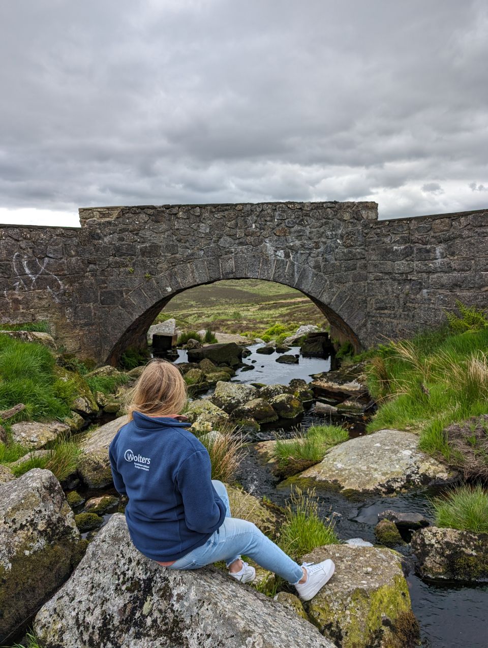 An einem Bachlauf sitzend blickt eine Frau auf eine alte Steinbrücke in den Wicklow Mountains in Irland. Die Szene fängt die ruhige und leicht melancholische Schönheit der rauen irischen Landschaft an diesem bekannten und romantischen Ort ein.