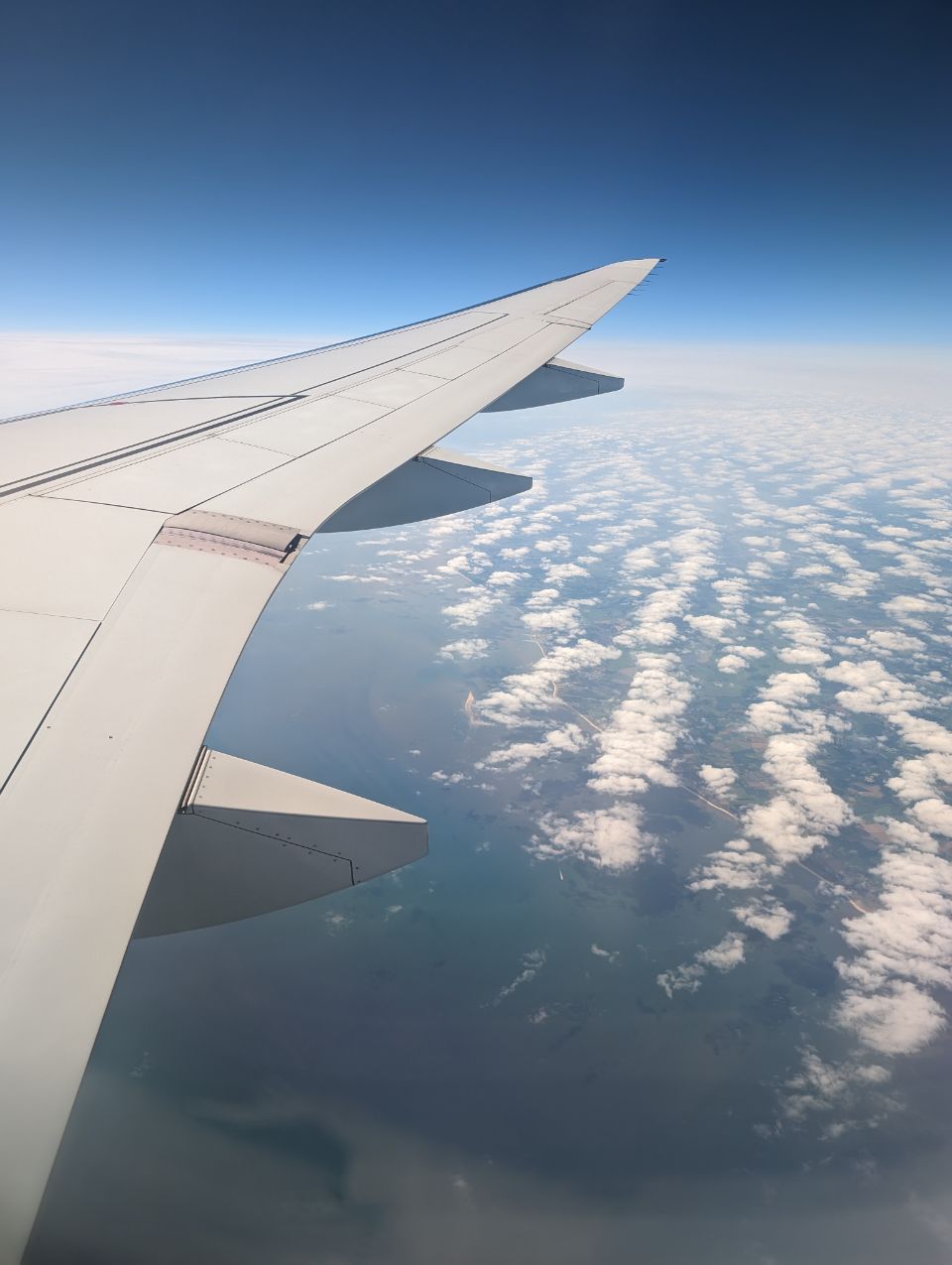 Hoch über den Wolken wurde dieser Blick aus einem Flugzeugfenster eingefangen. Man erkennt die Tragfläche, die über einem Feld aus Schönwetterwolken schwebt, während darunter die Küstenlinie von Land und Meer sichtbar wird, was eine friedliche Reise-Atmosphäre erzeugt.