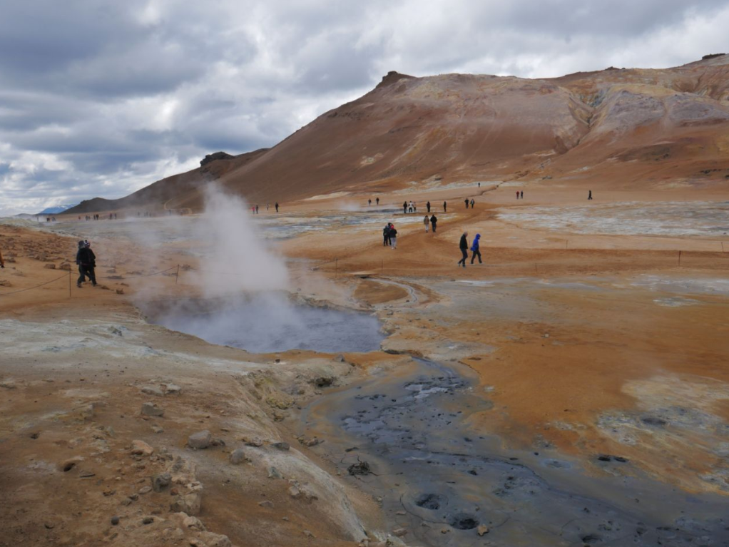 Auf diesem Bild ist die surreal wirkende Landschaft des Geothermalgebiets Hverir in Island zu sehen. Man erkennt die dampfenden Fumarolen und die durch Mineralien bunt gefärbte, karge Erde, durch die Besucher spazieren, um die vulkanischen Aktivitäten aus nächster Nähe zu erleben.