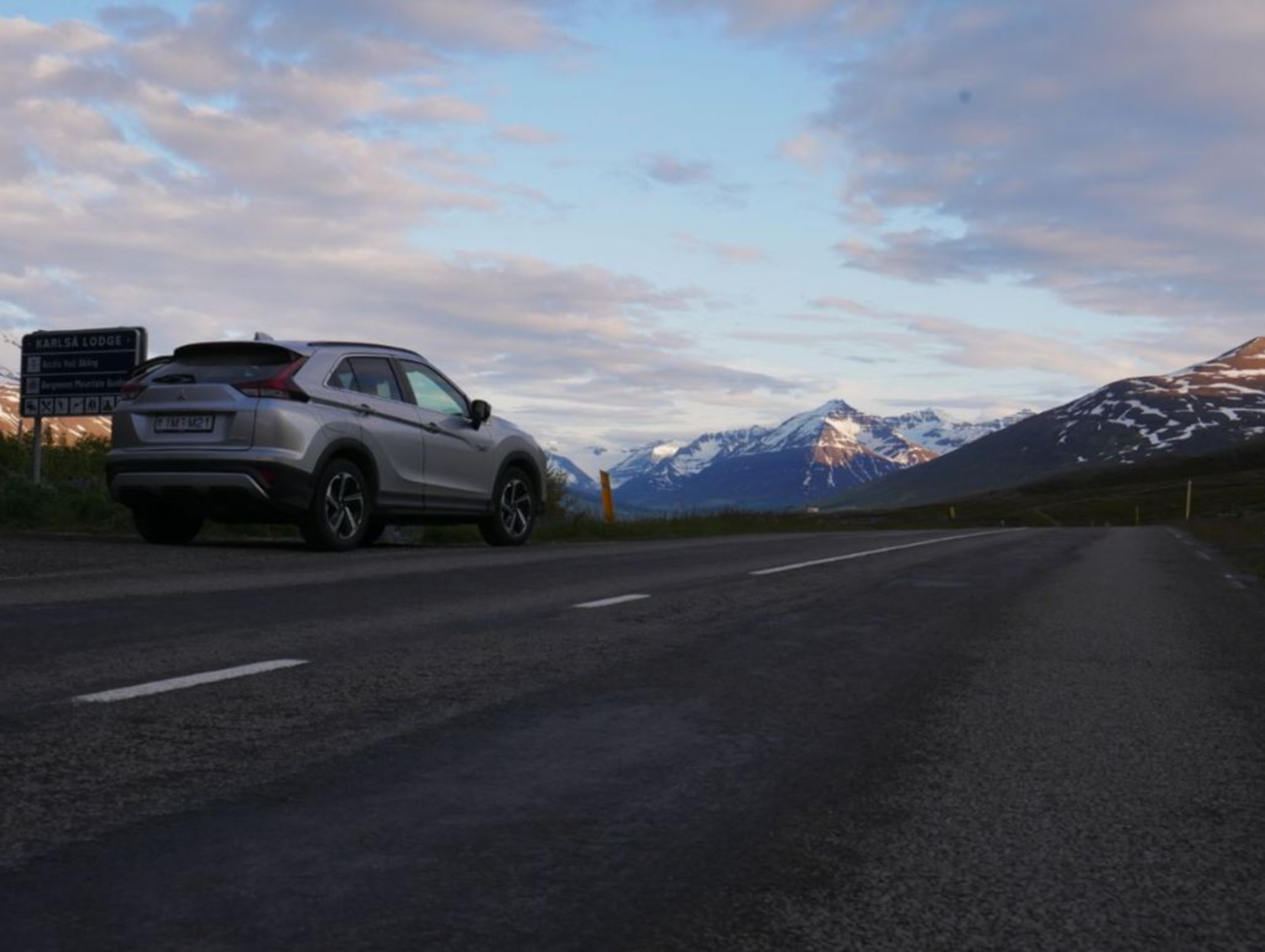Auf diesem Bild ist ein Auto zu sehen, das an einer Straße inmitten der beeindruckenden Berglandschaft von Nordisland geparkt ist. Die Aufnahme in der Abenddämmerung fängt mit den von der Sonne angestrahlten, schneebedeckten Gipfeln und der leeren Straße die Essenz eines Roadtrips durch die weite Natur Islands ein.