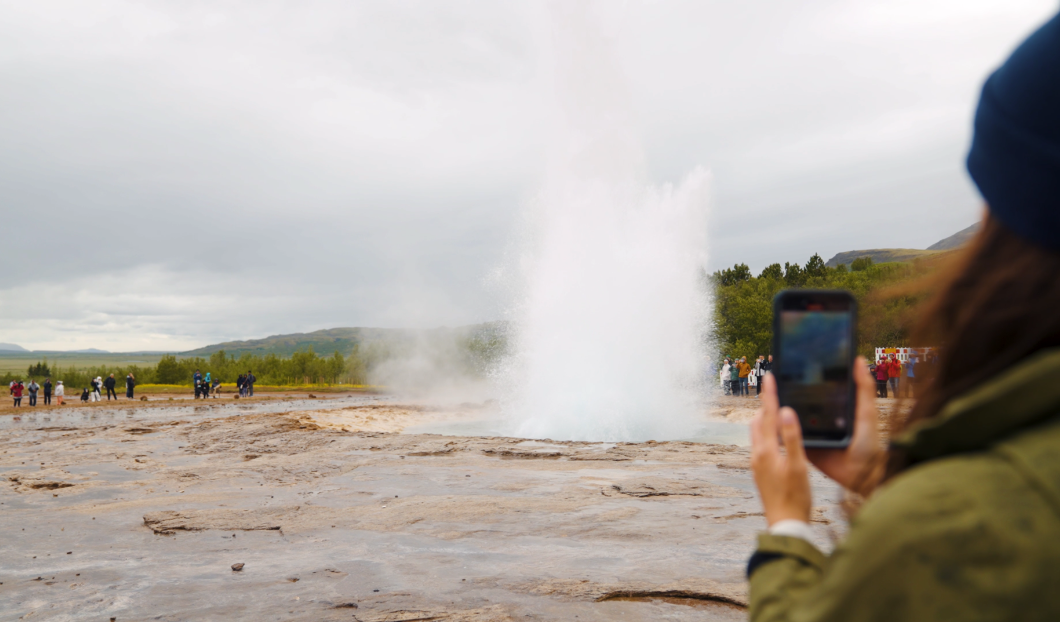 Den Ausbruch des Geysirs Strokkur in Island hält eine Person in diesem Bild mit ihrem Smartphone fest. Die Aufnahme zeigt die hohe Wasserfontäne inmitten des geothermalen Feldes, umgeben von zahlreichen Zuschauern, die dieses weltberühmte Naturspektakel beobachten.