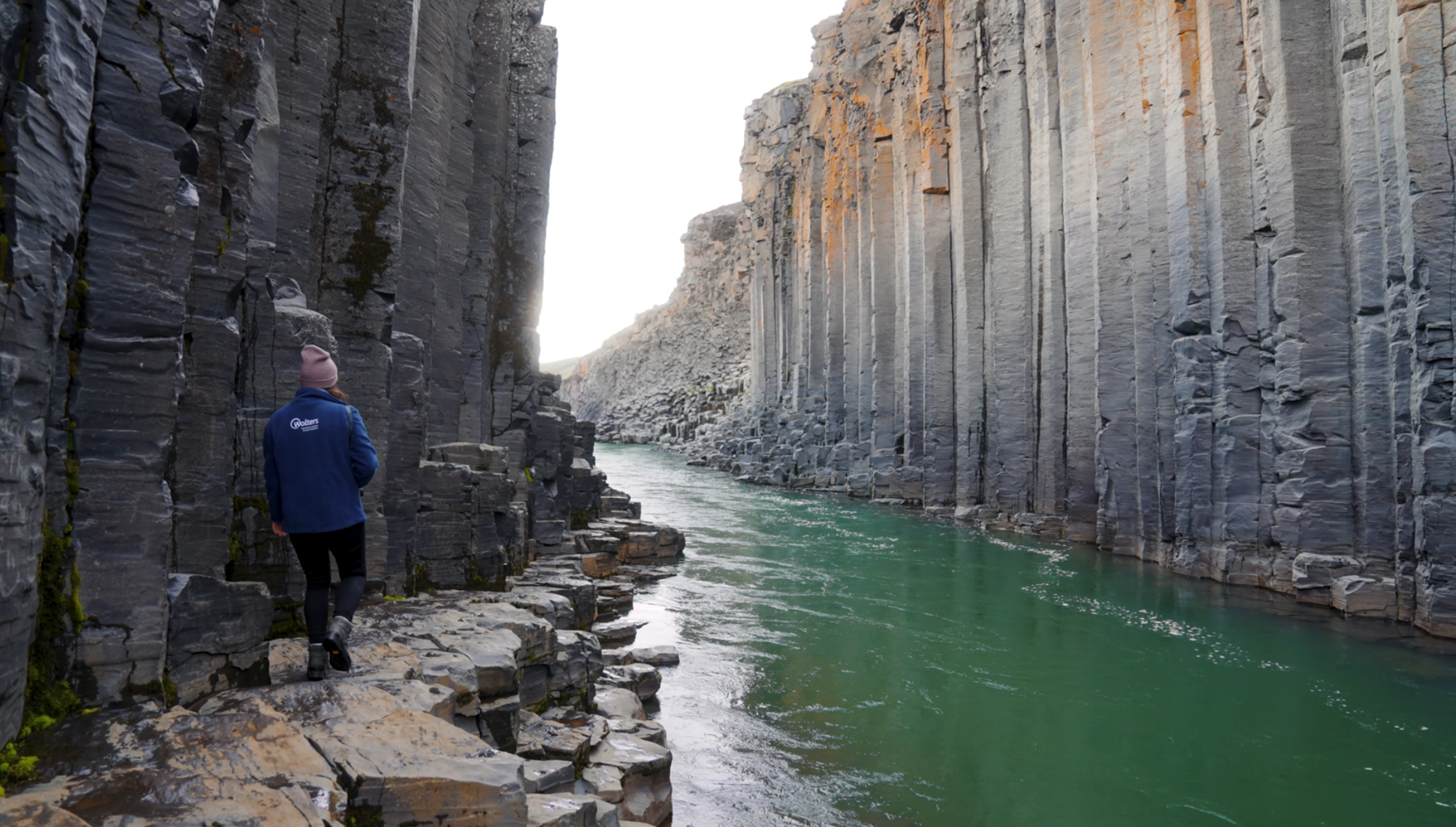 Umgeben von beeindruckenden, sechseckigen Basaltsäulen wandert eine Frau durch den Stuðlagil Canyon in Island. Die Aufnahme fängt die einzigartige Schönheit dieser geologischen Formation ein, bei der der türkisfarbene Fluss einen eindrucksvollen Kontrast zu den dunklen, ebenmäßigen Steinsäulen bildet.
