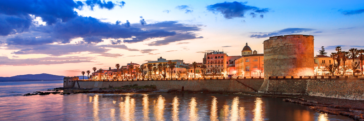 Panorama der Altstadt von Alghero auf Sardinien bei Sonnenuntergang, mit beleuchteter Stadtmauer und reflektierendem Licht im Meer.