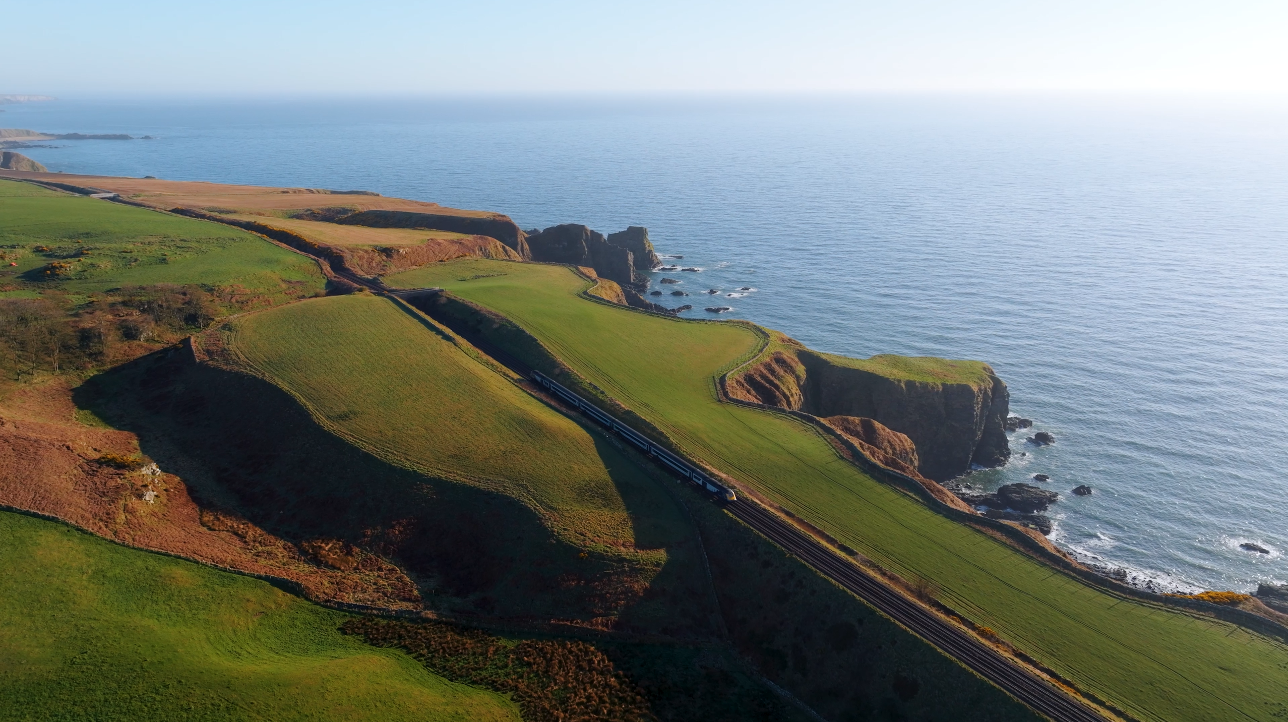 Luftaufnahme eines Zuges, der entlang der grünen Klippen über dem Meer fährt. Die Bahnlinie schlängelt sich durch die hügelige Küstenlandschaft Schottlands mit Blick auf die Nordsee. 