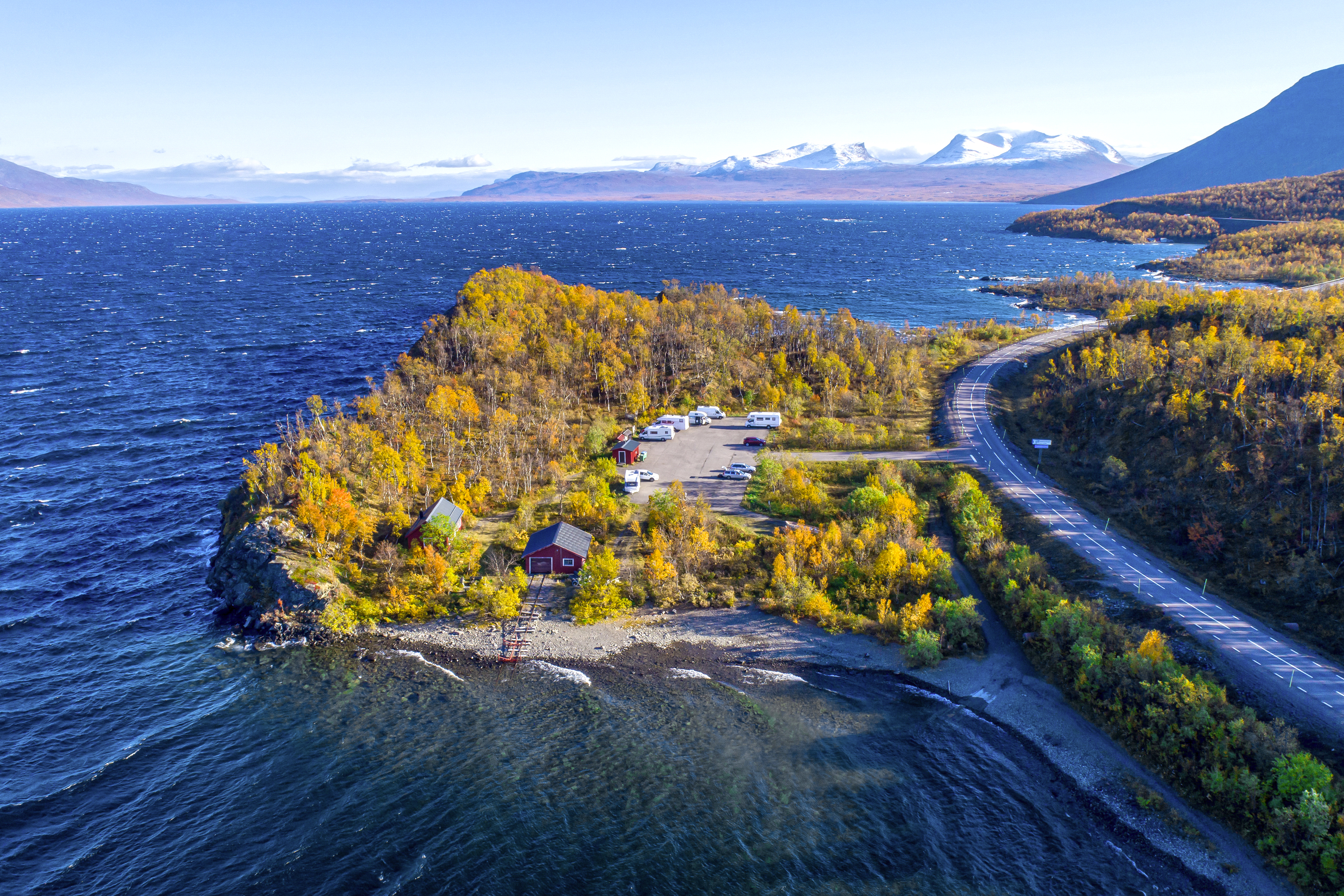 Panoramastraße am Torneträsk in Schwedisch-Lappland Straße schlängelt sich entlang des tiefblauen Torneträsk-Sees in Nordschweden. Herbstfarben leuchten, im Hintergrund liegen schneebedeckte Berge.