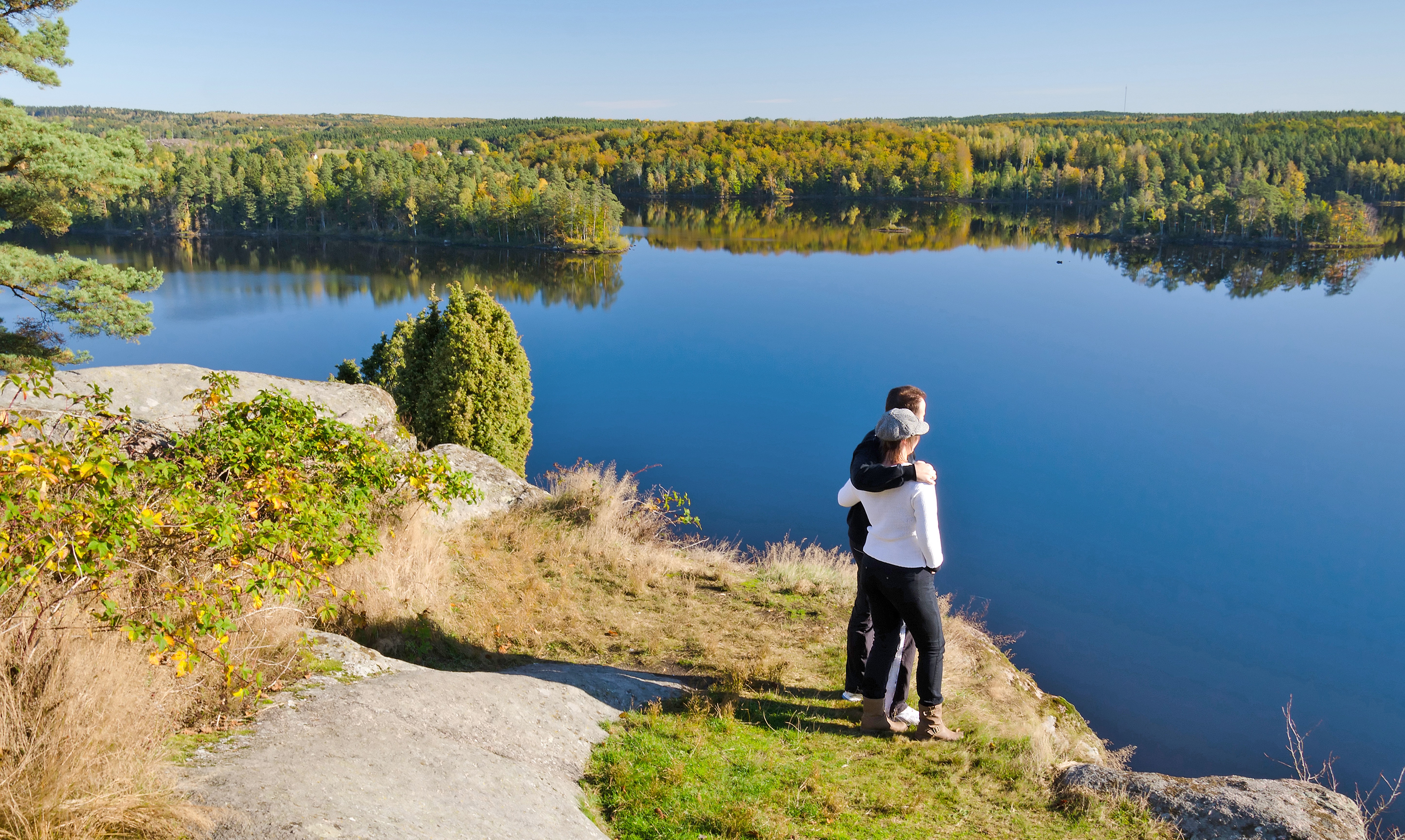 Pärchen am See in Südschweden Zwei Personen umarmen sich auf einem Felsen mit Blick auf einen stillen See, umgeben von Bäumen und blauem Himmel - friedliche Momentaufnahme in der Natur Schwedens.