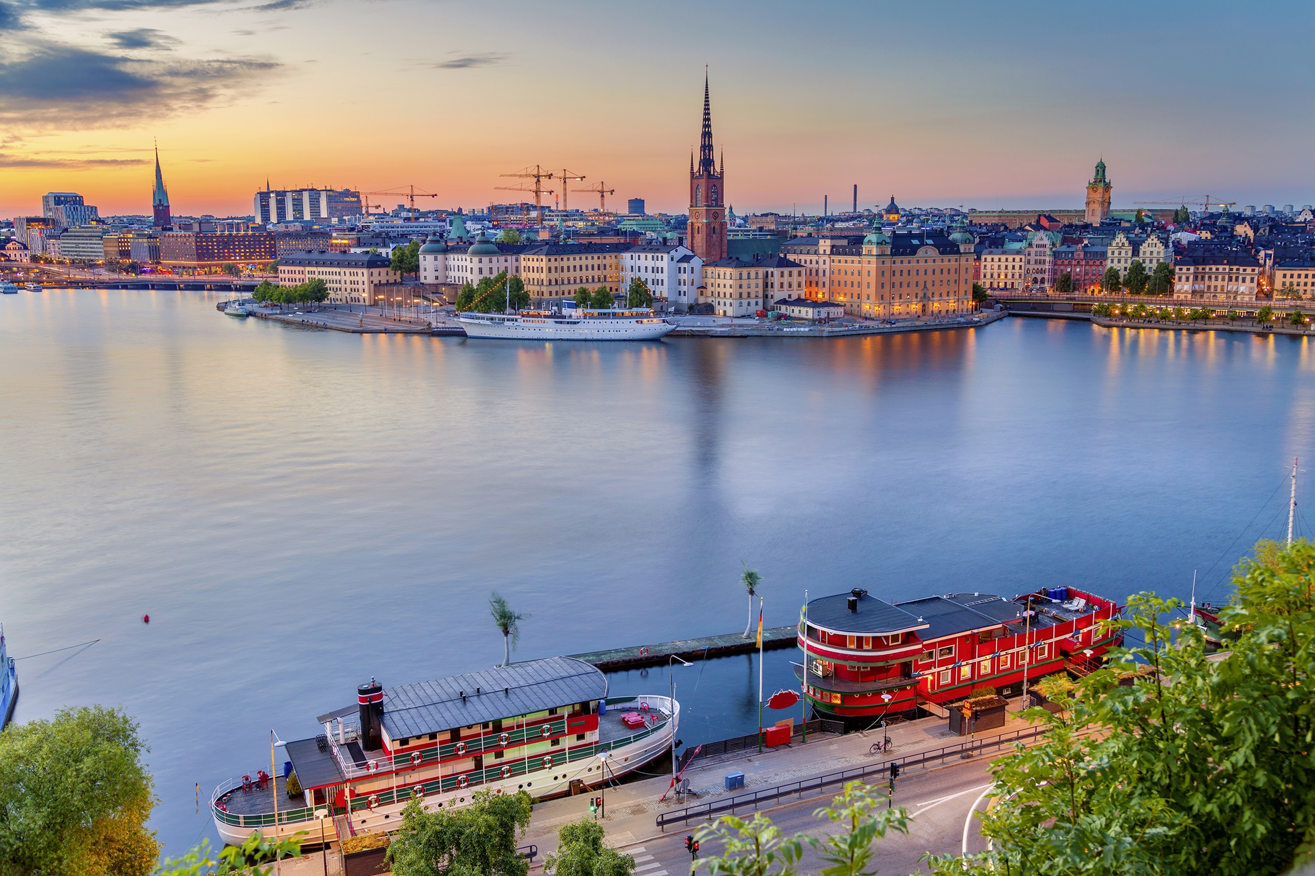 Stockholm Eine Luftaufnahme der Stadt Stockholm bei Sonnenuntergang mit Blick auf historische Gebäude, Kirchen und Boote im Hafen. Das Wasser spiegelt den farbenfrohen Himmel wider.