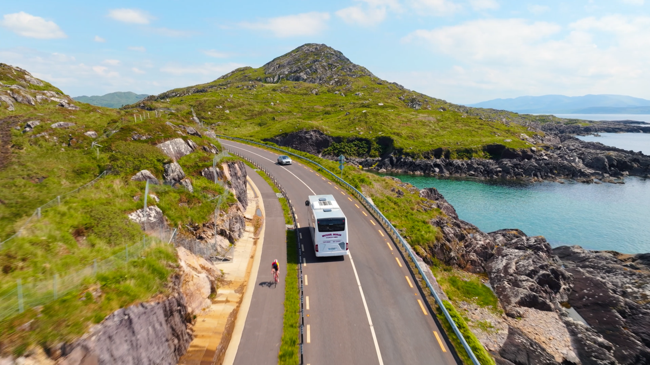 Auto und Reisebus auf der Küstenstraße des Ring of Kerry in Irland – beliebte Panoramaroute mit Meerblick und grünen Hügeln.