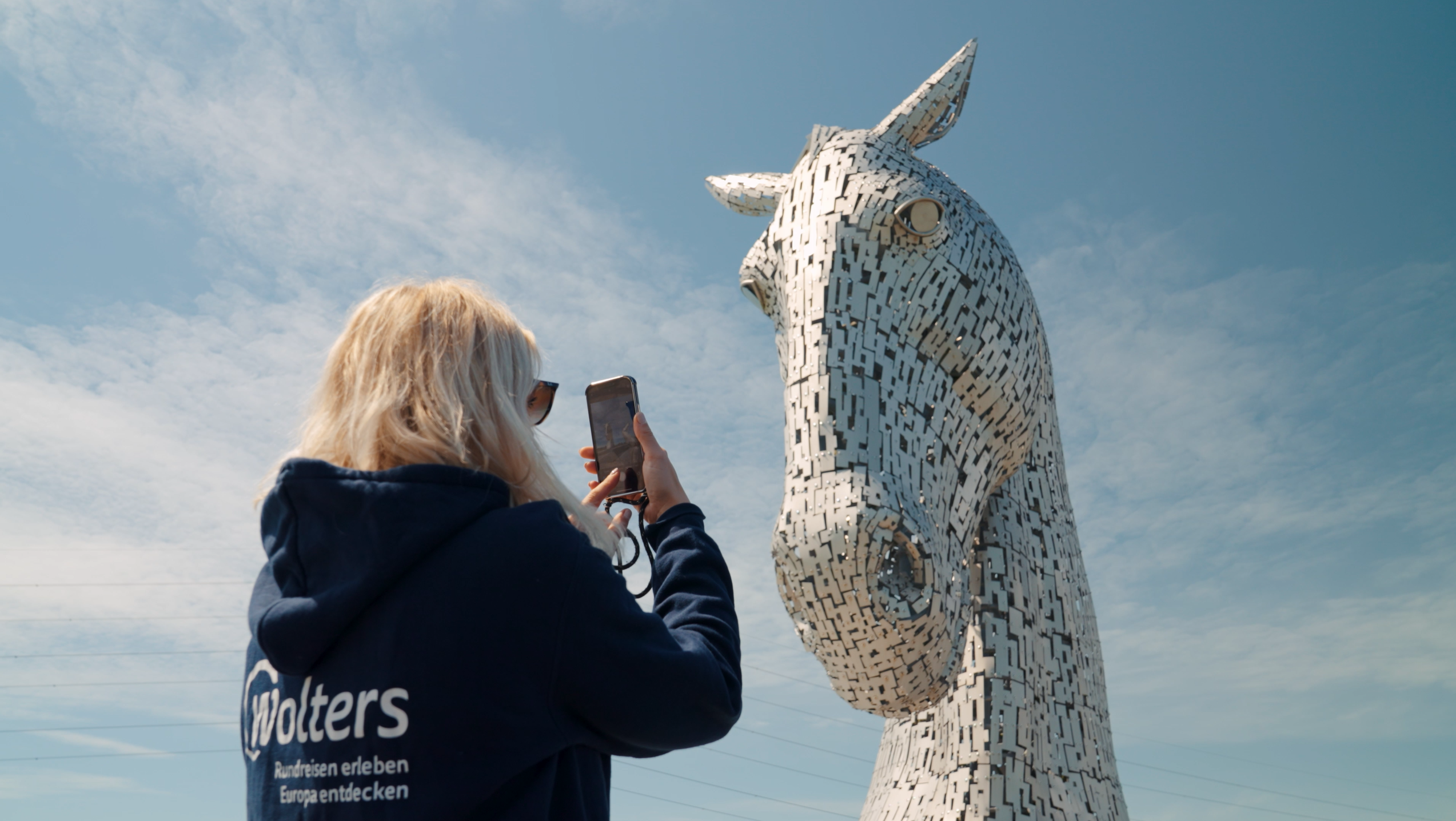 Eine Frau macht während ihrer Reise nach Schottland ein Foto von der beeindruckenden, aus Stahl gefertigten Pferdekopf-Skulptur der Kelpies in Falkirk, eine weltberühmte kulturelle Sehenswürdigkeit und ein Highlight für jeden Urlaub.