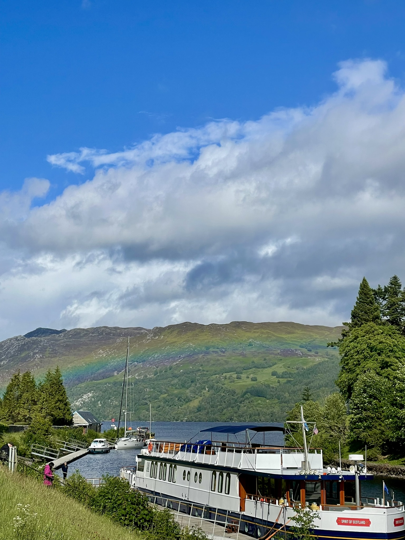 Ein Ausflugsboot liegt in einer Schleuse am berühmten Loch Ness in den schottischen Highlands, während sich ein leuchtender Regenbogen über die grünen Berge spannt und diese Reise durch die malerische Natur Schottlands zu einem unvergesslichen Urlaubserlebnis macht.