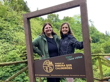 Gruppenreise auf den Azoren - Naturerlebnis und Gemeinschaft genießen Zwei Reisende posieren lächelnd in einem hölzernen Bilderrahmen mit der Aufschrift „Ribeira Grande – Salto do Cabrito“ auf den Azoren. Umgeben von üppiger grüner Vegetation zeigt das Foto die Freude am gemeinsamen Reisen und Erkunden der Natur.