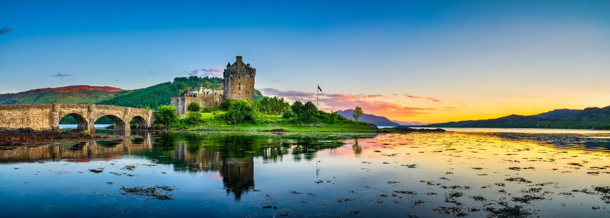 Das mittelalterliche Eilean Donan Castle in Schottland mit Steinbrücke, umgeben von Wasser und Bergen, im goldenen Licht des Sonnenuntergangs. Eine der bekanntesten Sehenswürdigkeiten der schottischen Highlands.