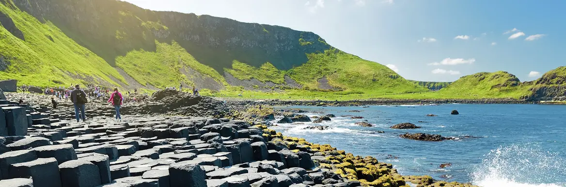 Das Bild zeigt den Giant's Causeway in Nordirland. Die Basaltsäulen werden von Touristen begangen und rechts sieht man das Meer, was mit Gischt an die Ufer schlägt. Im Hintergrund sind grüne Berge. 