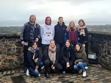 Gruppenreise nach Edinburgh – gemeinsames Erlebnis in Schottlands Hauptstadt Eine Gruppe von Reisenden steht lachend auf einer alten Steinmauer mit Blick über die Stadt Edinburgh in Schottland. Umgeben von herbstlicher Landschaft vermittelt das Foto die Freude am gemeinsamen Reisen und Erkunden kultureller Highlights während einer Gruppenreise nach Großbritannien.