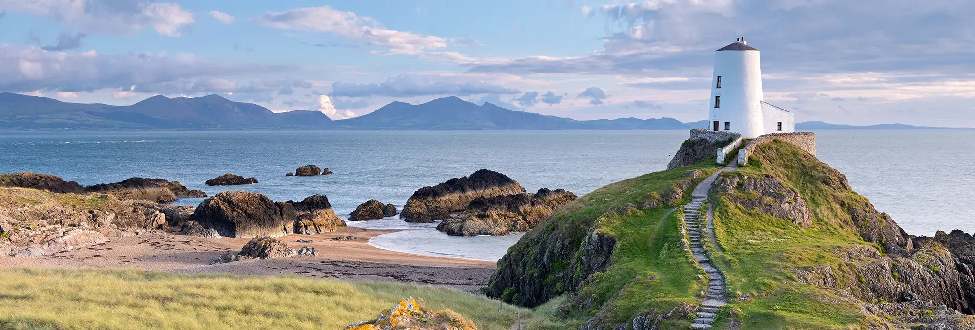 Auf dem Bild sieht man einen weißen Leuchtturm an der Küste. Er liegt auf einem grasbewachsenem Hügel und ein steinerner Weg führt hoch. Im Hintergrund ist das Meer und weitere Berge am Horizont.