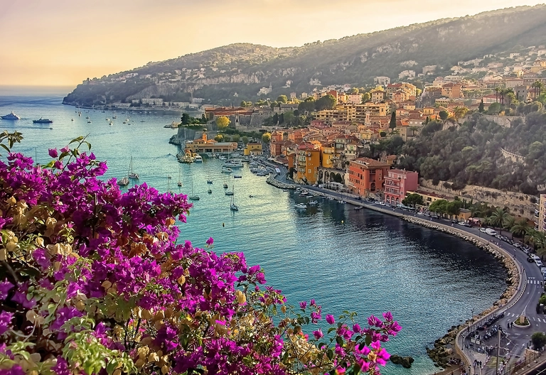 Blick auf die Küstenstadt Villefranche-sur-Mer an der französischen Riviera mit bunten Häusern, Booten im Hafen und blühendem Bougainvillea im Vordergrund.