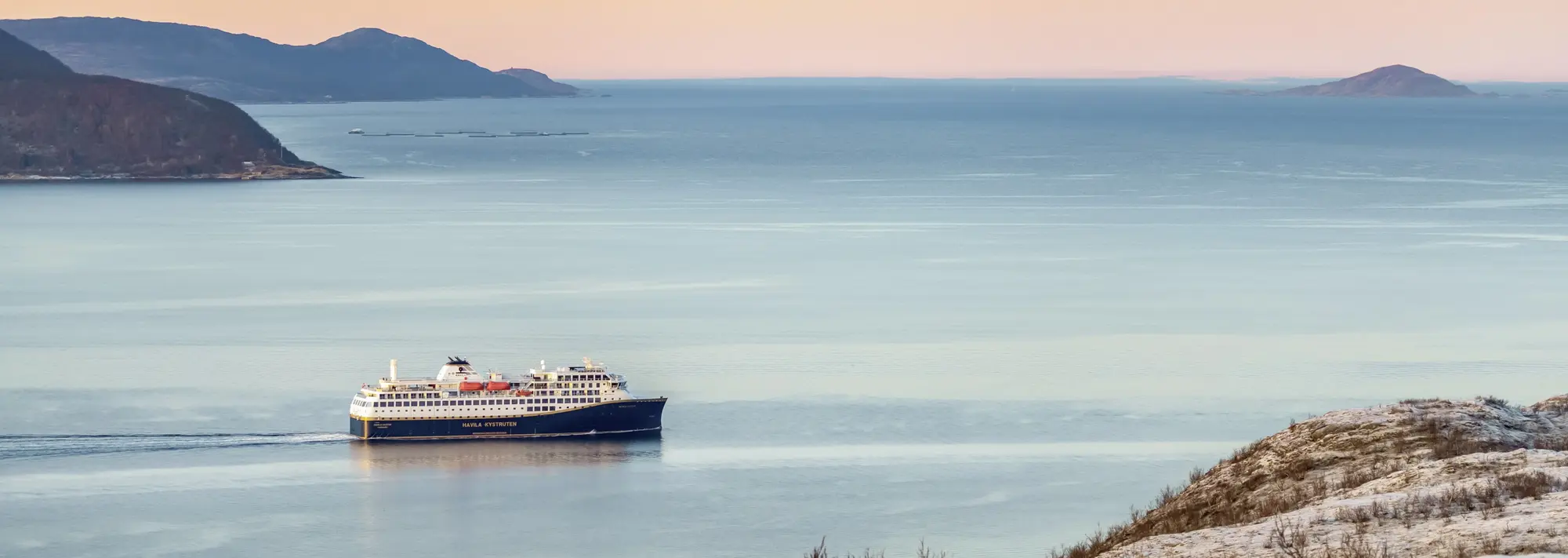 Ein modernes Postschiff der Havila Kystruten gleitet bei Sonnenuntergang durch die ruhigen Gewässer der norwegischen Fjordlandschaft und bietet Passagieren auf dieser unvergesslichen Reise einen malerischen Anblick auf die Küste.