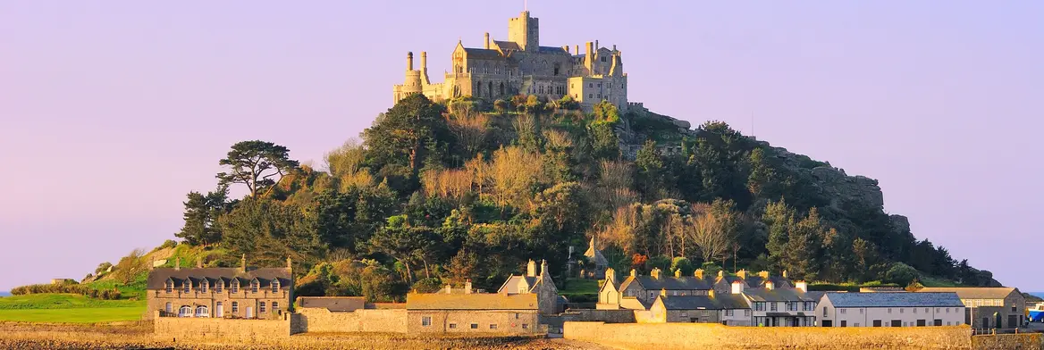 Die Gezeiteninsel St. Michael’s Mount in Cornwall mit ihrer imposanten Burganlage auf einem bewaldeten Hügel, umgeben von kleinen Häusern und dem Meer im warmen Abendlicht.