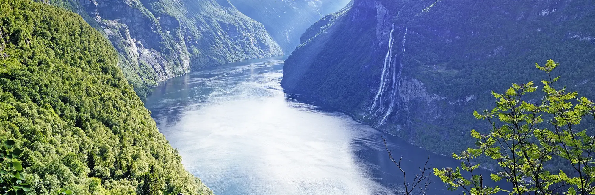 Das Bild zeigt den berühmten Geirangerfjord in Norwegen, umgeben von steilen, grünen Berghängen. Im Hintergrund stürzt der Wasserfall "Sieben Schwester" ins Tal. Diese spektakuläre Fjordlandschaft gehört zum UNESCO-Weltnaturerbe und ist ein Highlight jeder Norwegen-Rundreise.