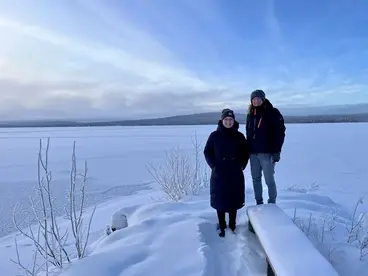 Winter in Lappland - Reisende genießen die verschneite Landschaft am gefrorenen See Zwei Reisende stehen warm eingepackt auf einem schneebedeckten Steg vor einem zugefrorenen See in Lappland. Im Hintergrund erstreckt sich eine weite Winterlandschaft unter blauem Himmel - ein typisches Bild für die klare, friedliche Atmosphäre des Nordens während einer Lapplandreise.