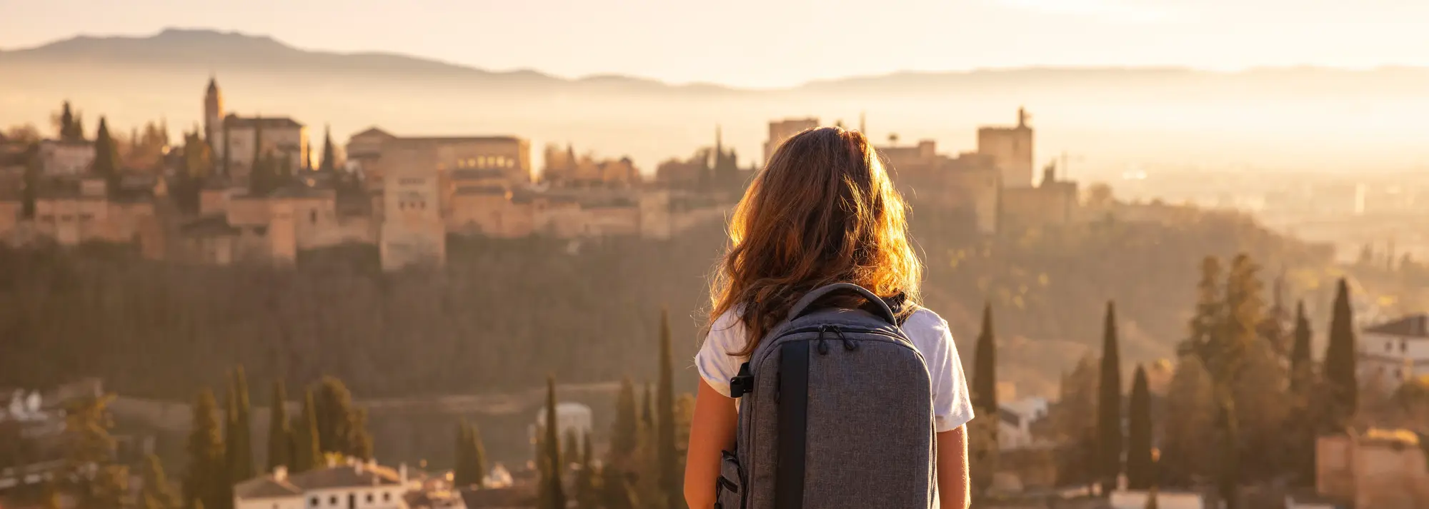 Eine Reisende mit Rucksack blickt bei Sonnenuntergang auf die Stadt Granada und die Alhambra in Andalusien. Das warme Licht taucht die historische Kulisse in goldene Töne und vermittelt die Atmosphäre einer kulturellen Reise durch Südspanien - perfekt für Urlauber, die Geschichte, Architektur und authentische Erlebnisse lieben.